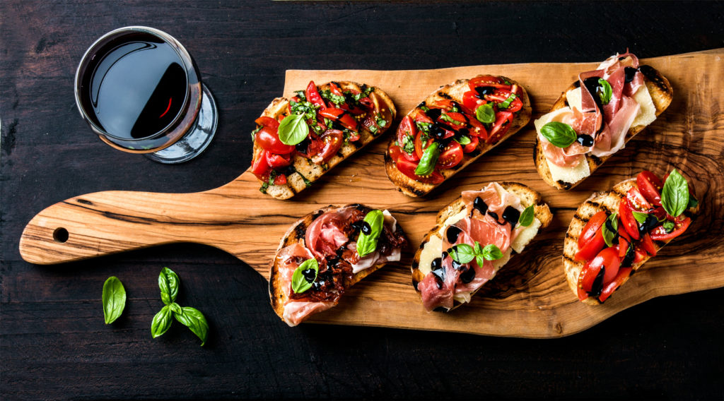 Bruschetta set and glass of red wine. Small sandwiches with Brushetta set for wine. Variety of small saBruschetta sandwiches with prosciutto, tomatoes, parmesan cheese, fresh basil and balsamic creme served with glass of red wine on rustic wooden board over dark background, top view